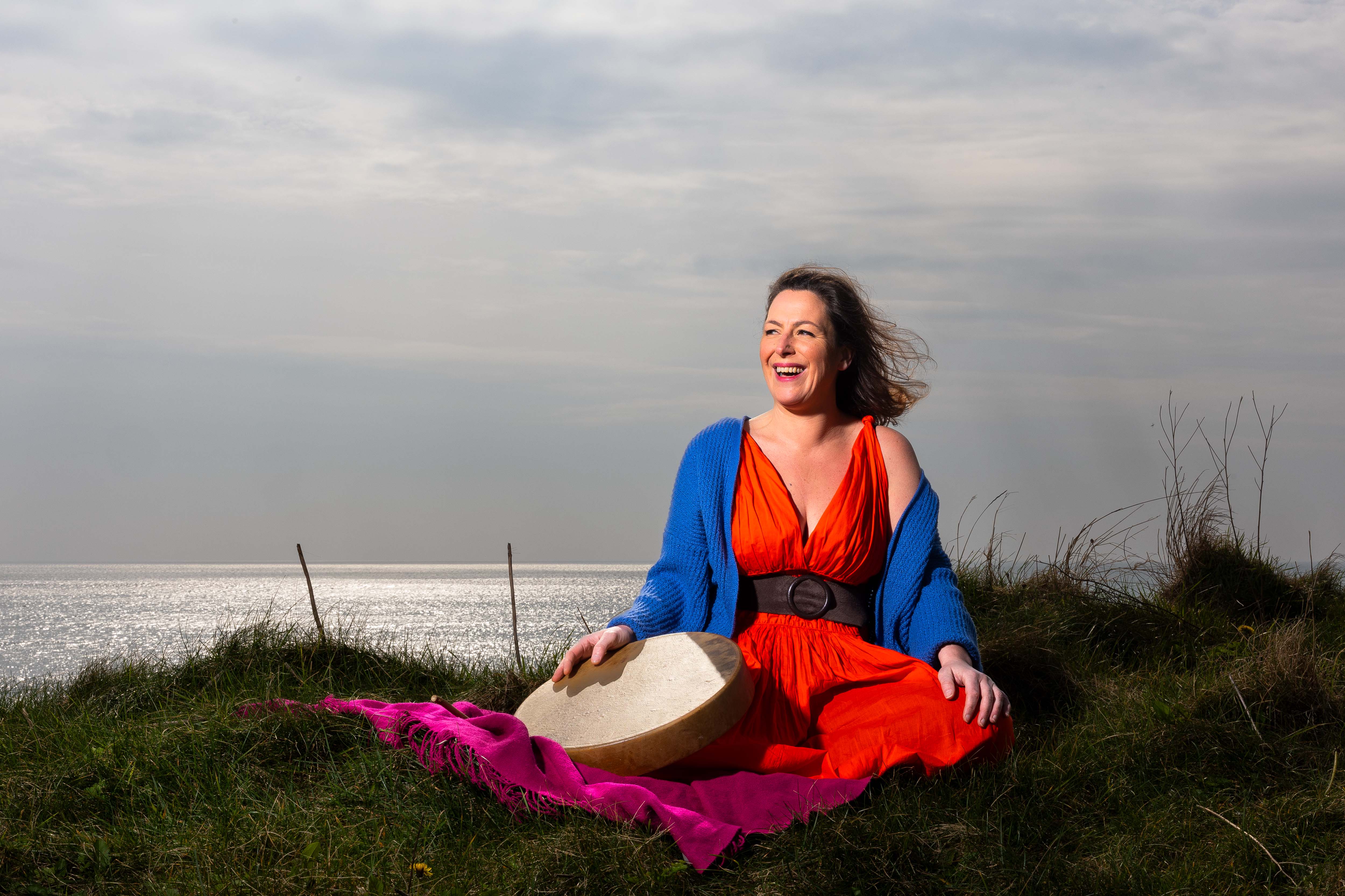 Ciara McDonnell sitting in the grass by the sea, holding a bodhrán drum, laughing in the wind